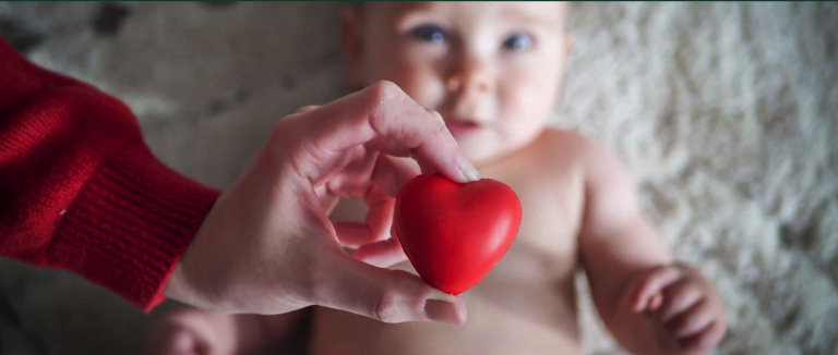 A baby lays down with a person's hand holding a rubber heart over it.